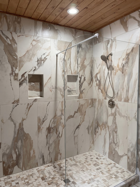 A modern walk-in shower stall with beige and brown marble-look wall tiles, a matching mosaic tile floor, a clear glass door, and a wood-paneled ceiling with a recessed light.