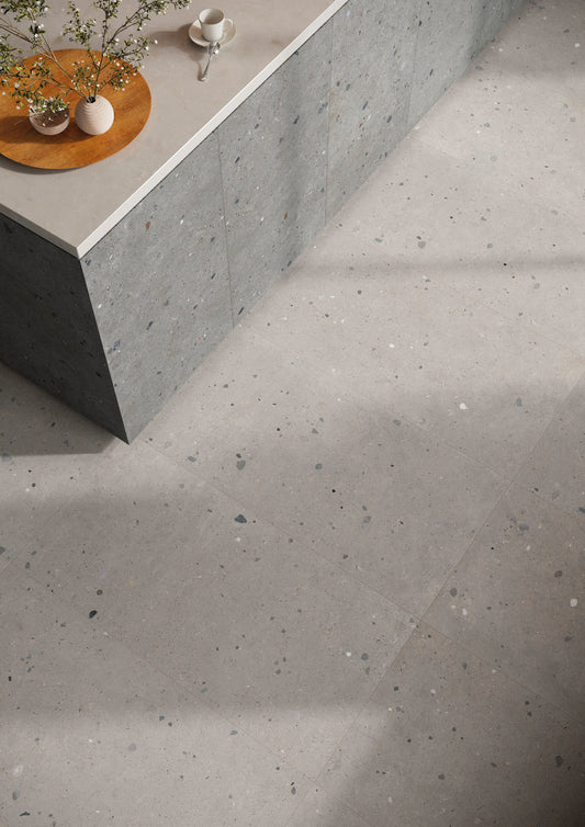 A high-angle view of a modern interior space showcasing large-format light gray concrete-look floor tiles with subtle dark and white speckles. A matching speckled stone countertop and dark gray concrete side panel are visible in the upper left, styled with a small wooden tray, vases, and a teacup.