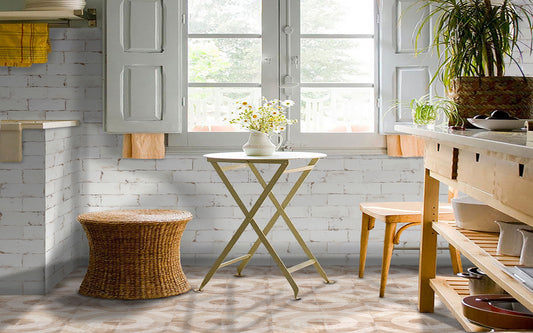A bright, styled interior corner with a small round folding table in the center, a woven wicker stool, a wooden counter with shelves, and a large potted plant near the window.