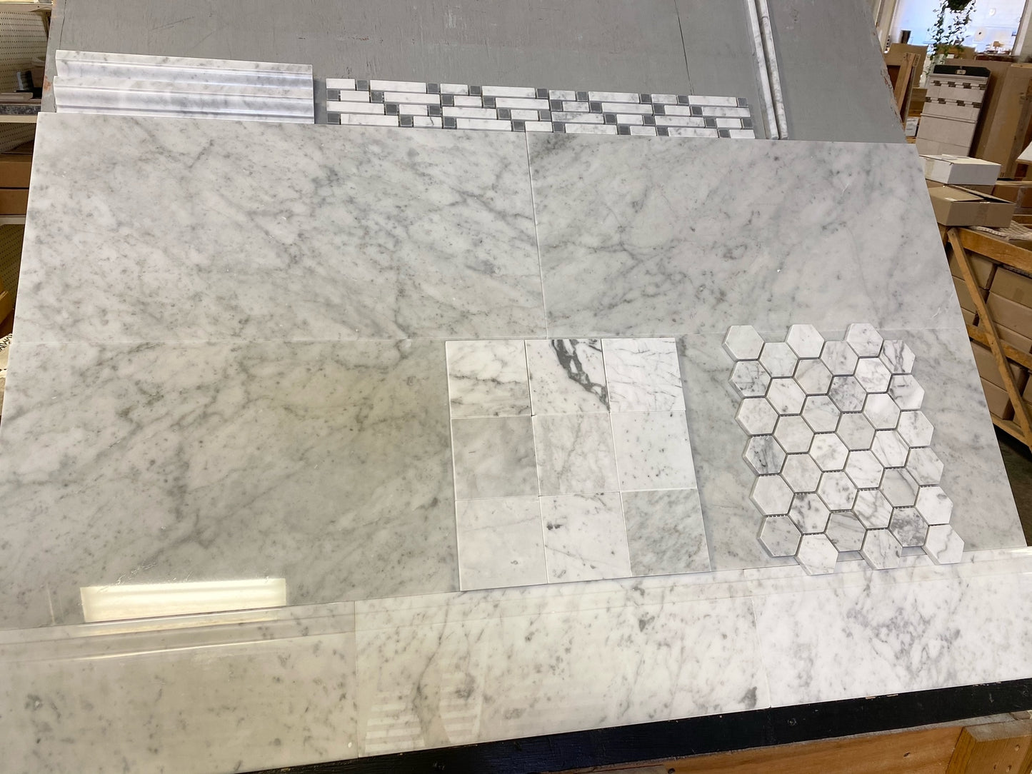 A large display of white marble tiles and mosaic samples, including a hexagonal tile pattern, arranged on a workbench in a tile showroom.