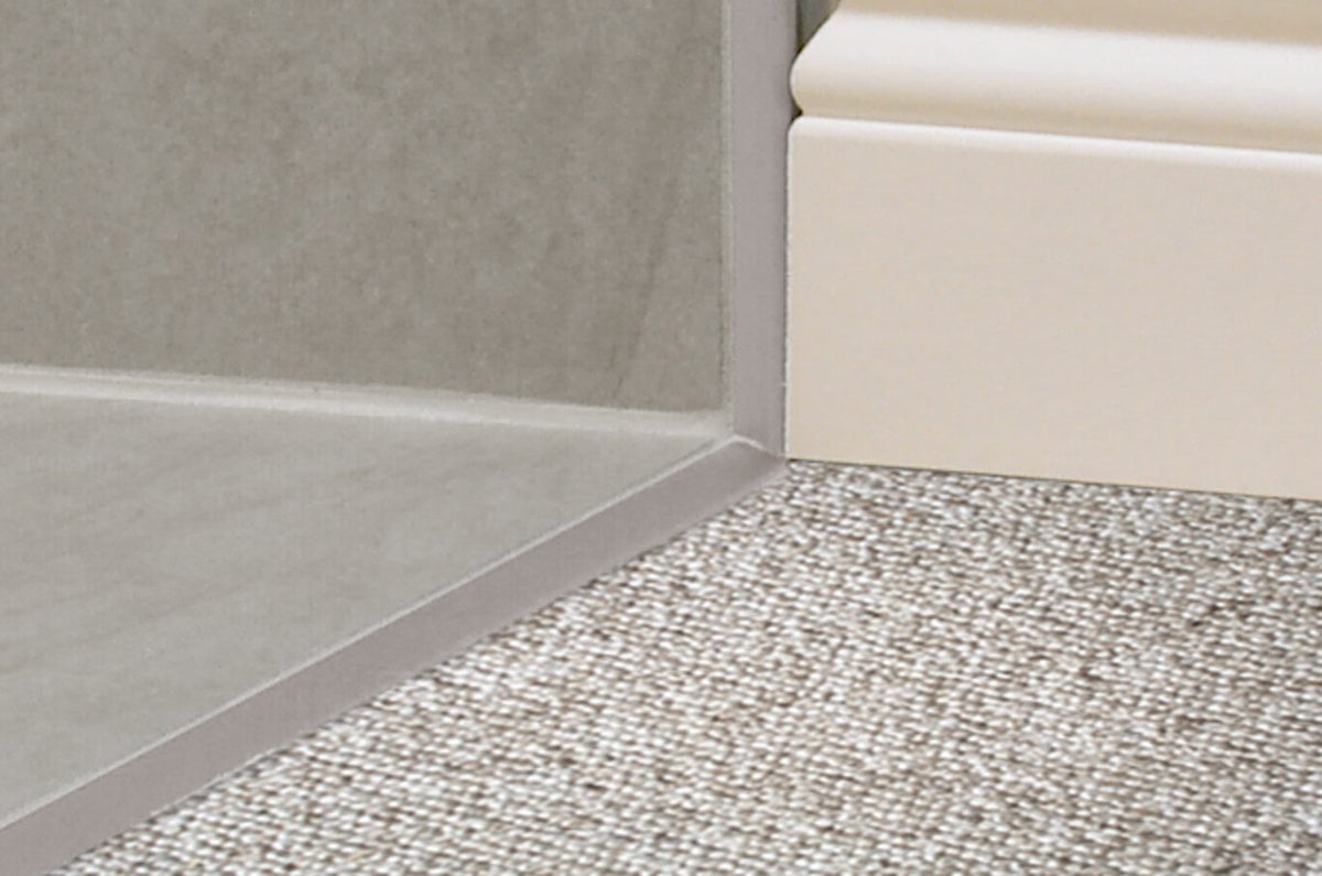 Close-up of a shower base corner with a textured beige and white pebble tile floor, a light gray tiled wall, and a cream-colored baseboard.