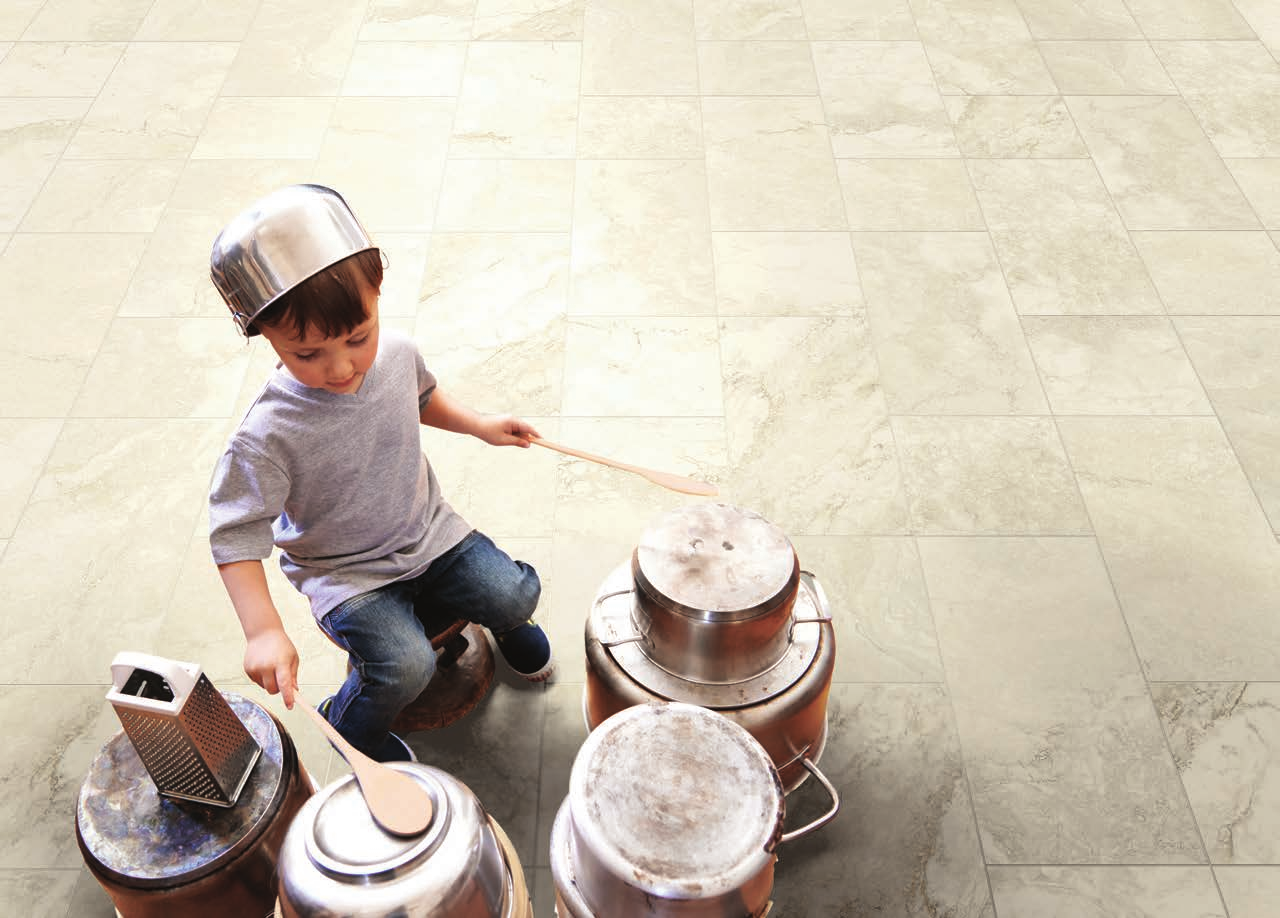 A young child wearing a metal pot as a helmet plays with metal cans and wooden spoons on a light beige marble tile floor.