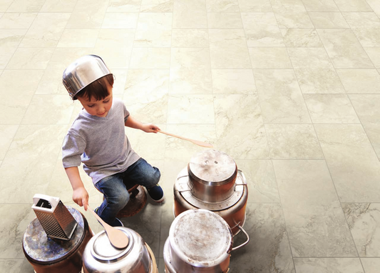 A young child wearing a metal pot as a helmet plays with metal cans and wooden spoons on a light beige marble tile floor.