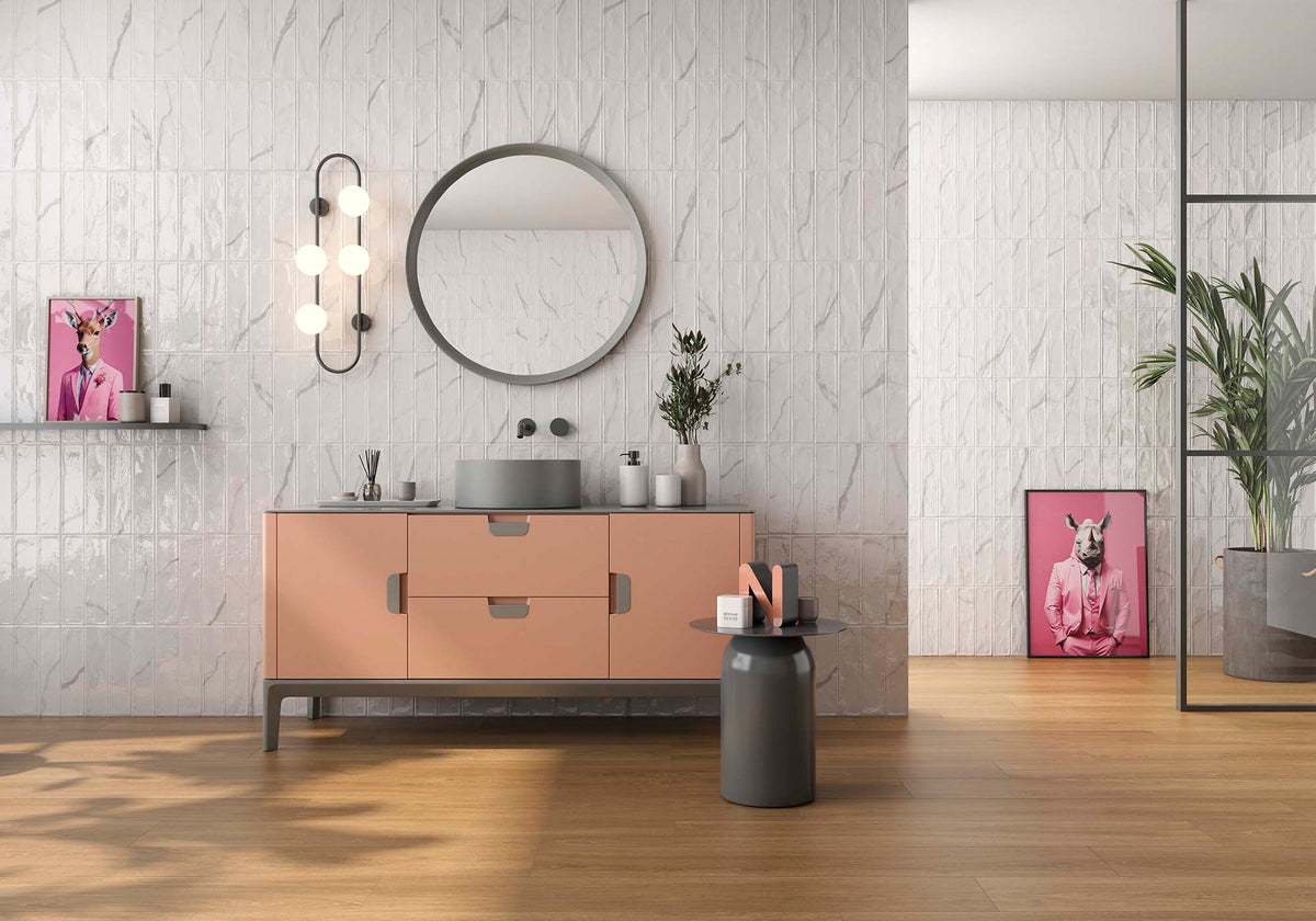 A modern bathroom vanity setup with a blush pink cabinet, a round backless mirror, a wall-mounted triple-bulb light, and a gray vessel sink, shown in a styled interior with wood flooring and pink framed art.