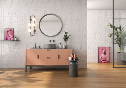 A modern bathroom vanity setup with a blush pink cabinet, a round backless mirror, a wall-mounted triple-bulb light, and a gray vessel sink, shown in a styled interior with wood flooring and pink framed art.