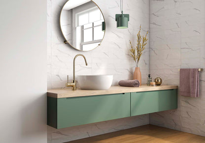 A modern bathroom vanity setup with a floating sage green cabinet, a light wood countertop, a white vessel sink, a gold faucet, a round wall mirror, and a hanging green pendant light. A towel is visible on the right side against white marble-look tiled walls.