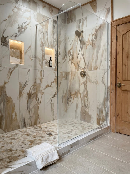 A modern walk-in shower enclosure with a clear glass door and curved glass panel, set against marble-look tiled walls and a mosaic tile floor. A white towel rests on the shower base, with a wooden door visible on the right.