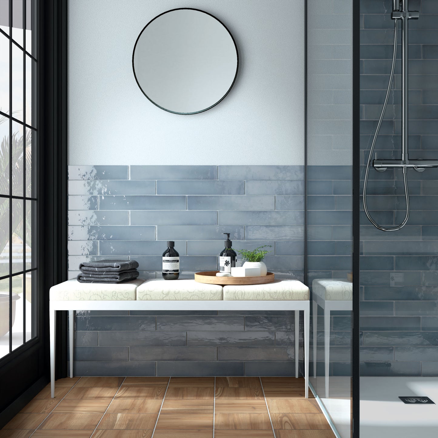 A modern bathroom interior with a white bench, blue-gray tiled wall, round black-framed mirror, wood floor, and a glass shower enclosure on the right.