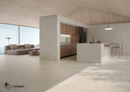A modern open-plan interior with a concrete kitchen island, wood cabinetry, bar stools, a large sofa in the living area, and a bright floor-to-ceiling window. The InTech+ logo appears in the lower-left corner.