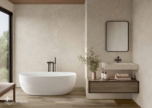 A modern bathroom interior featuring a white freestanding bathtub, a floating vanity with a white sink and mirror, wood flooring, and textured beige walls. A small window on the left shows greenery outside.