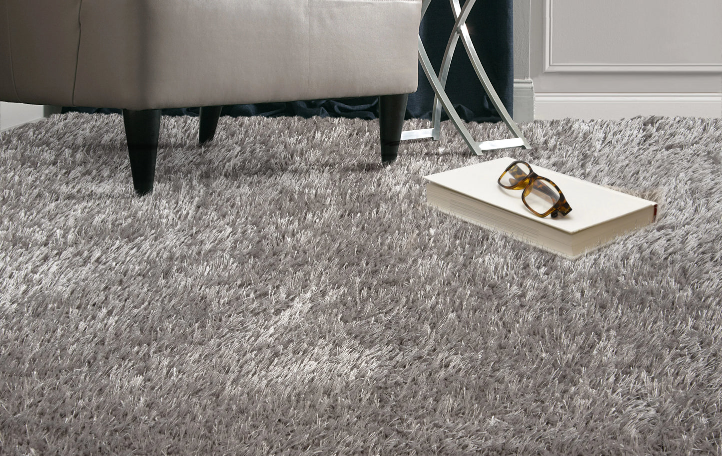 A gray shag area rug in a modern room setting, shown under a light gray upholstered ottoman with a book and eyeglasses on top.