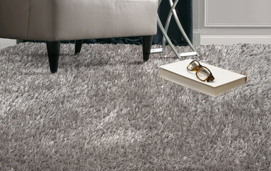 A gray shag area rug in a modern room setting, shown under a light gray upholstered ottoman with a book and eyeglasses on top.