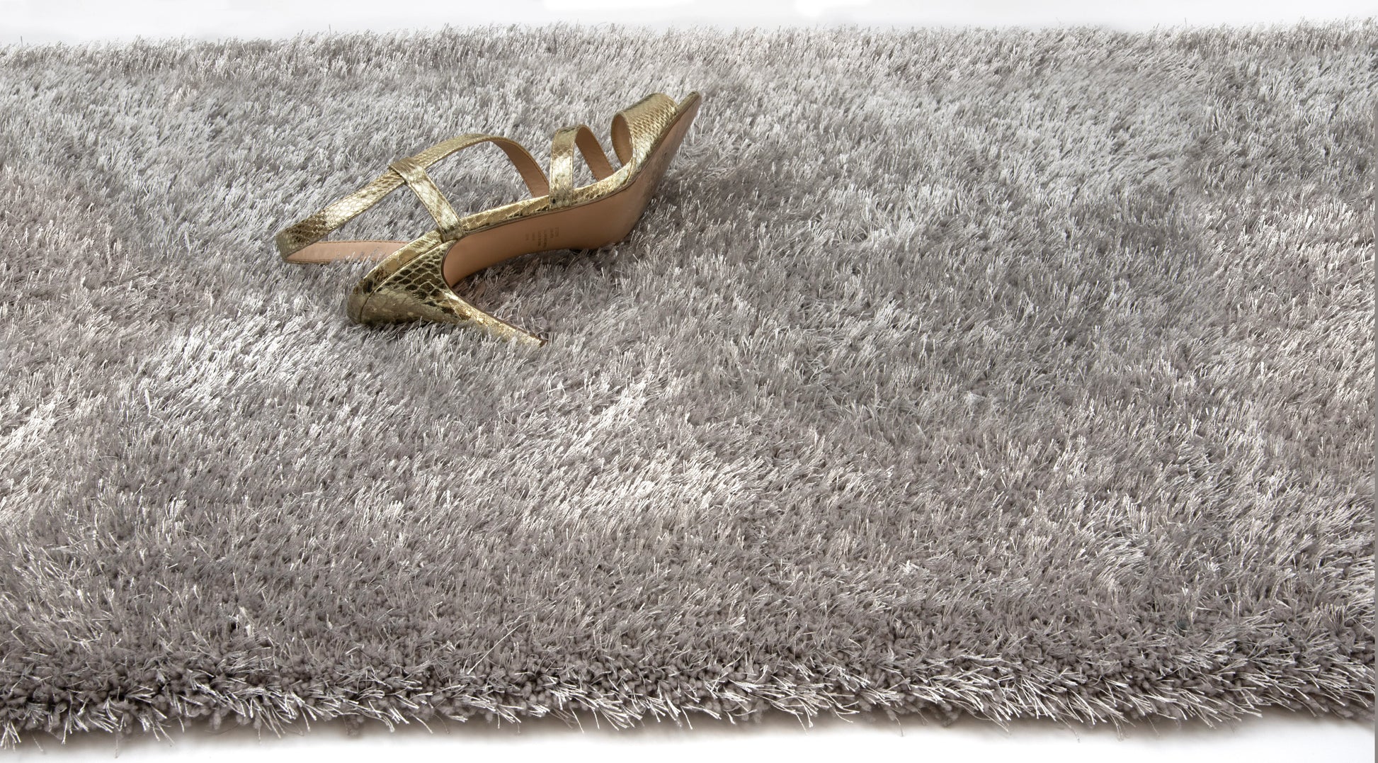 A single gold strappy high-heeled shoe placed on a thick, shaggy gray rug against a white background.