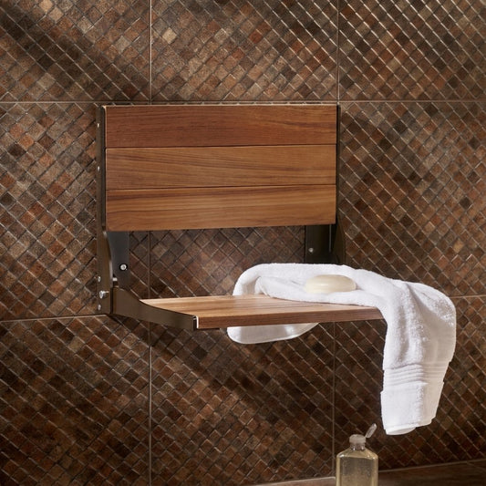 A wall-mounted wooden fold-down shelf with a backrest, shown installed on a brown mosaic tile wall. A white towel and a bar of soap are draped over the shelf.