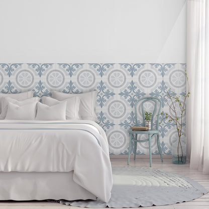 A styled bedroom scene featuring a decorative wallpaper with a blue and white geometric tile pattern on the lower half of the wall, paired with white bedding, a light blue vintage chair, and a white curtain.