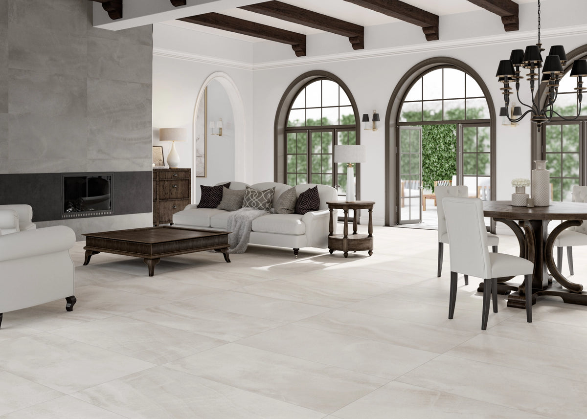 A large, bright living room with light beige stone-look floor tiles, featuring a white sofa, dark wood coffee table, dining set, arched windows, and dark wood ceiling beams.