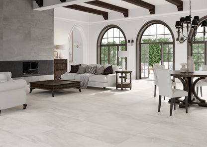 A large, bright living room with light beige stone-look floor tiles, featuring a white sofa, dark wood coffee table, dining set, arched windows, and dark wood ceiling beams.