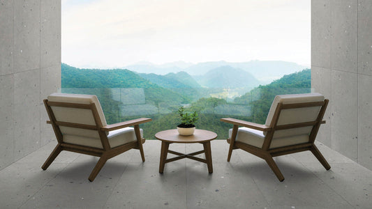 Two mid-century modern wooden lounge chairs with cream cushions and a small round wooden coffee table in between, set on a concrete patio with a scenic mountain landscape visible through a large glass window.