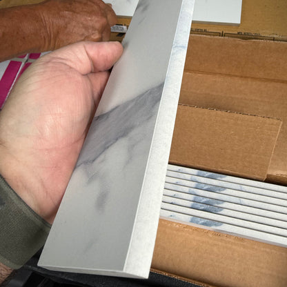 Hand holding a piece of white tile trim with a cardboard box of trim in the background