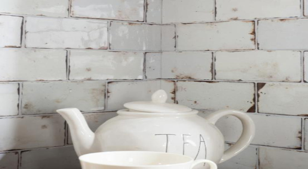 A white ceramic teapot with the word 'TEA' printed on the side, shown with a matching white cup in front of a distressed white subway tile backsplash.