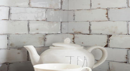 A white ceramic teapot with the word 'TEA' printed on the side, shown with a matching white cup in front of a distressed white subway tile backsplash.