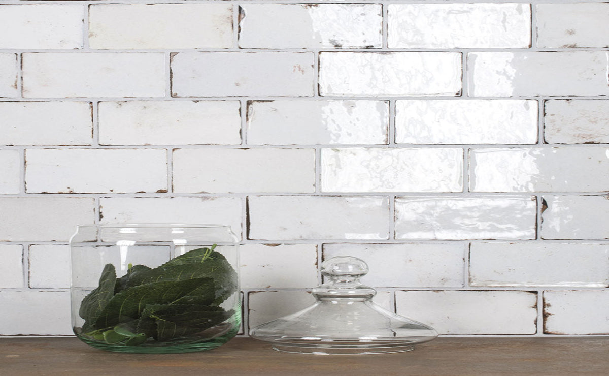 A clear glass jar filled with green leaves sits on a wooden surface in front of a white subway tile wall, with a matching clear glass lid beside it.