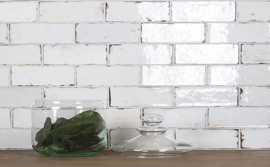 A clear glass jar filled with green leaves sits on a wooden surface in front of a white subway tile wall, with a matching clear glass lid beside it.