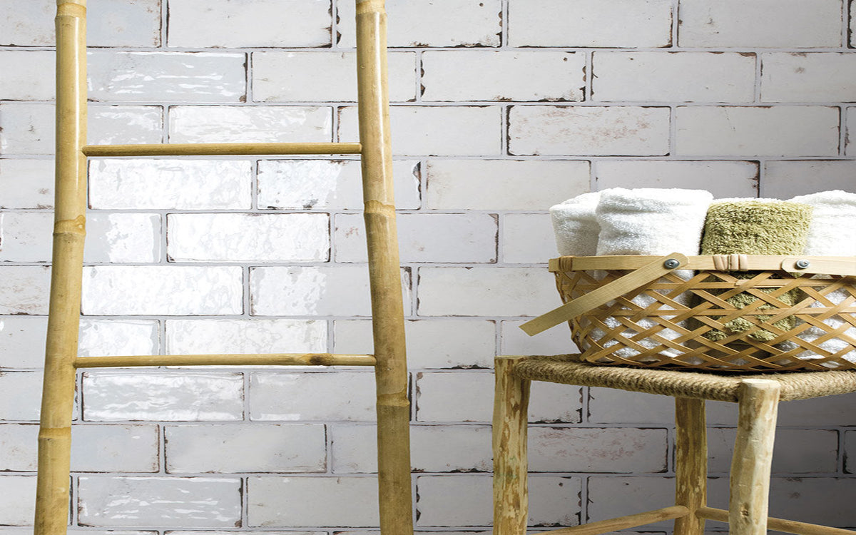 A natural bamboo ladder leans against a distressed white brick wall next to a woven rattan stool holding a basket of folded white towels.