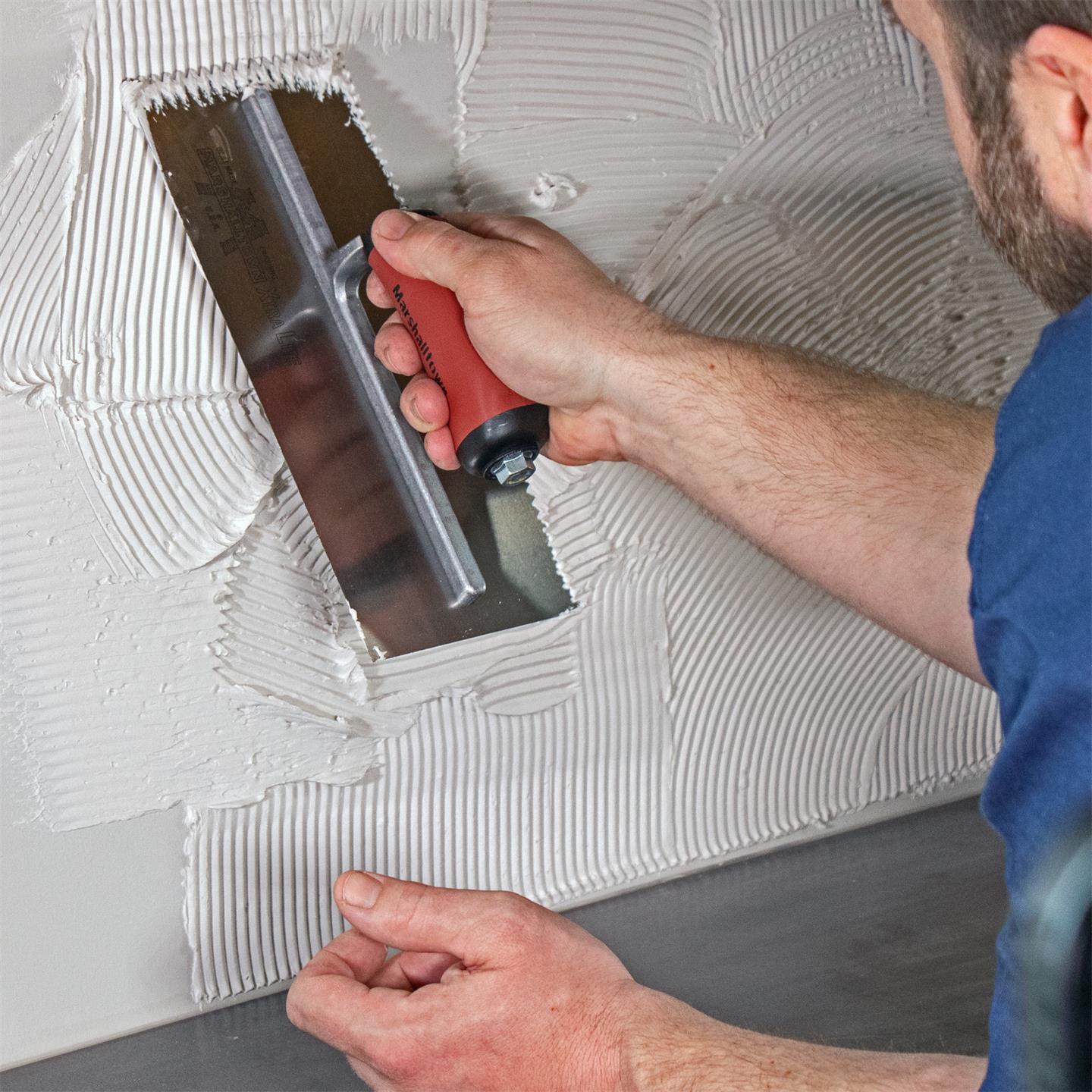 Tile installer with blue shirt using a V-Shaped Notched Trowel with an maroon handle and a silver blade, spreading white mastic on a backsplash over a countertop.