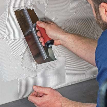 Tile installer with blue shirt using a V-Shaped Notched Trowel with an maroon handle and a silver blade, spreading white mastic on a backsplash over a countertop.