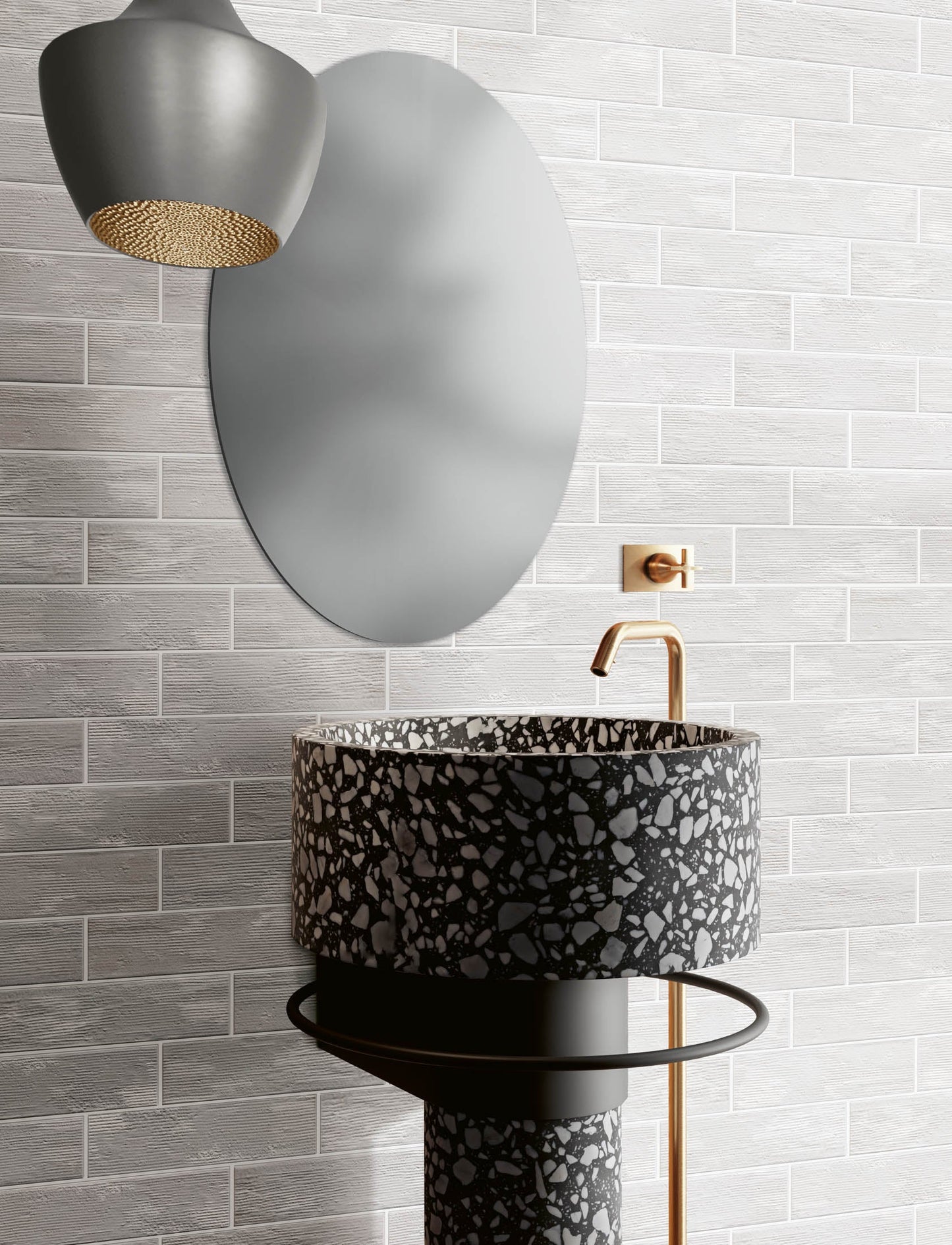 A modern bathroom scene featuring a round black and gray terrazzo vessel sink on a pedestal base, paired with a matte gold faucet, a large oval wall mirror, and a textured gray subway tile wall.