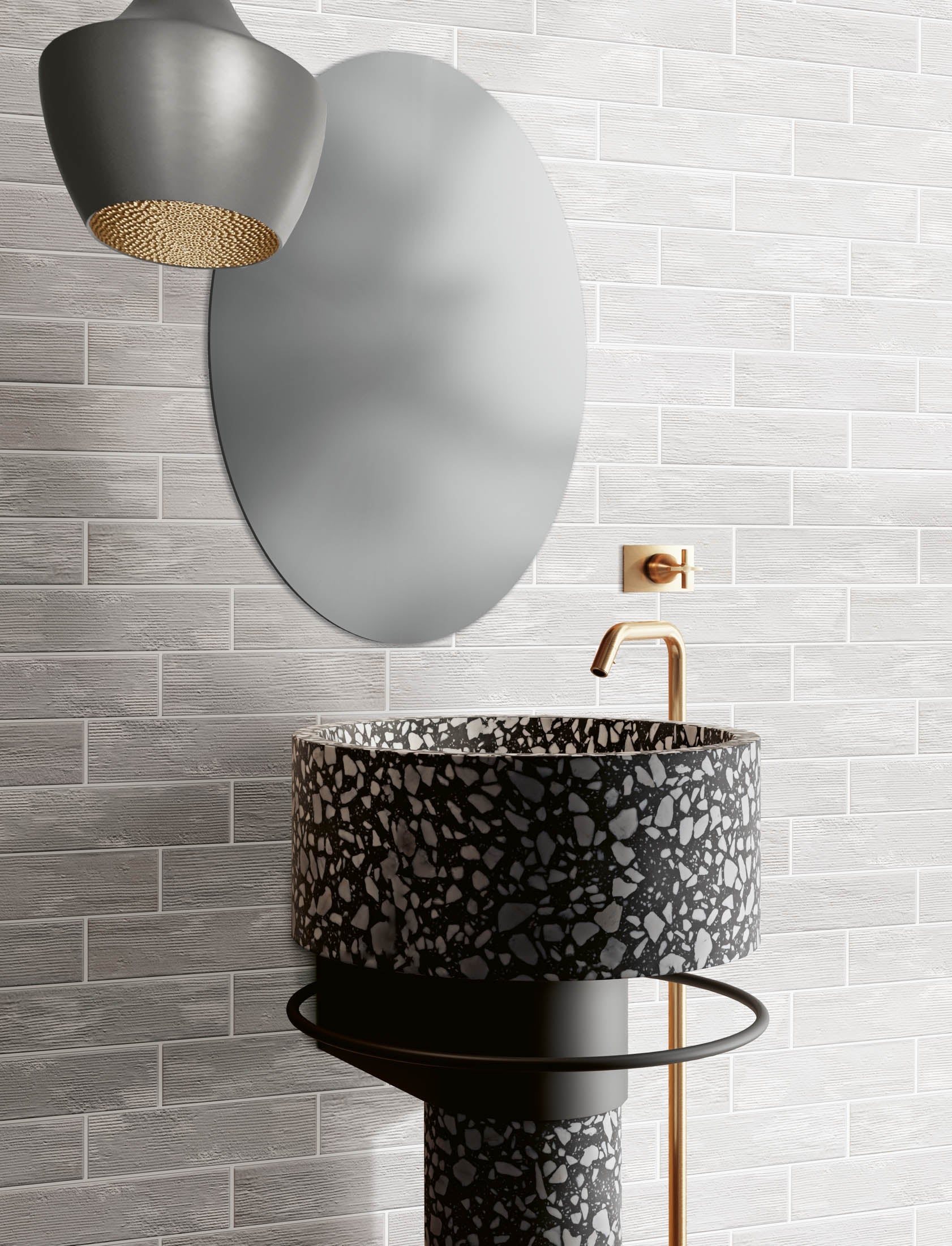 A modern bathroom scene featuring a round black and gray terrazzo vessel sink on a pedestal base, paired with a matte gold faucet, a large oval wall mirror, and a textured gray subway tile wall.