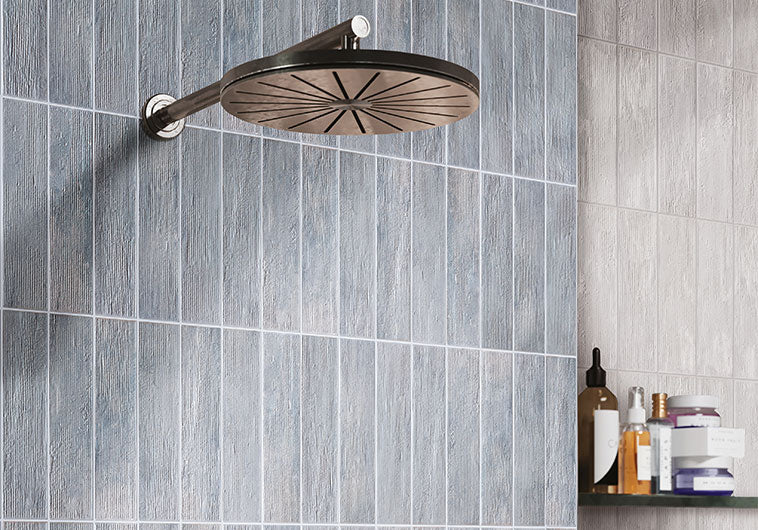 A round wall-mounted shower head with a bronze-finished frame and starburst pattern, mounted on a gray wood-look tiled wall in a bathroom setting.